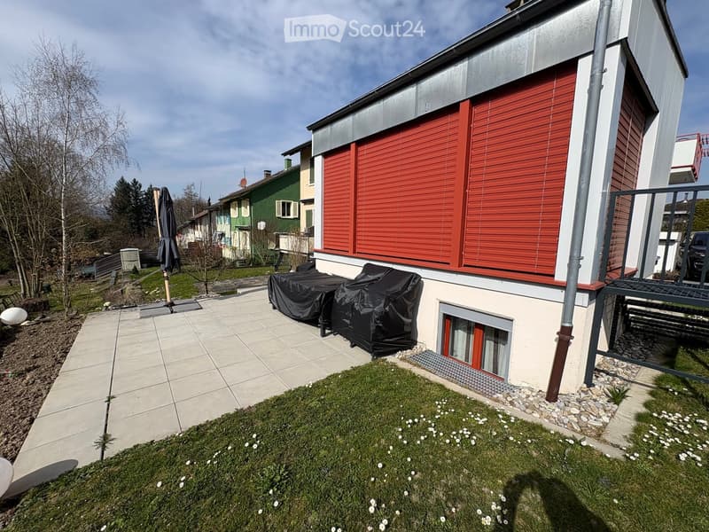 Modern house exterior with a raised ground floor, patio area, covered outdoor furniture, and a well-maintained lawn.