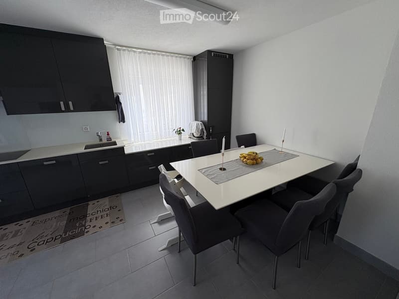 Modern kitchen and dining area with dark grey cabinets, white dining table, six chairs, and a small sink area with a faucet and a window with white curtains.