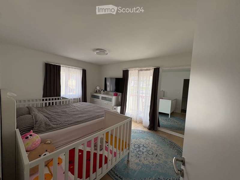 A well-lit bedroom with a large bed, a nightstand, a mirror, and a television. The room features a round rug and a crib with toys.