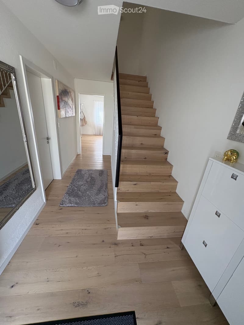 A modern hallway with wooden flooring, a staircase leading upwards, and a small grey rug placed near the stairs. The walls are painted white, and there are mirrors and a small cabinet visible on the right side.