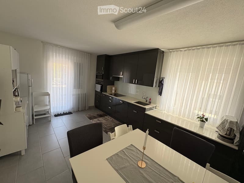 Modern kitchen with dark cabinets, white countertops, a dining area with a table and chairs, and a small bar area with a stool. The space features large windows with sheer curtains and a tiled floor.