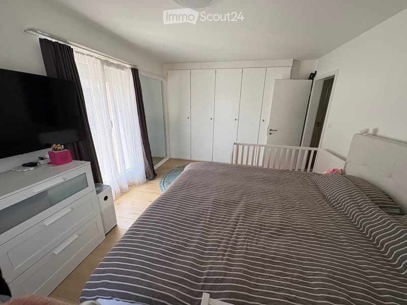Modern bedroom with a large bed, striped bedding, white wardrobe, TV on a stand, and large windows with curtains.