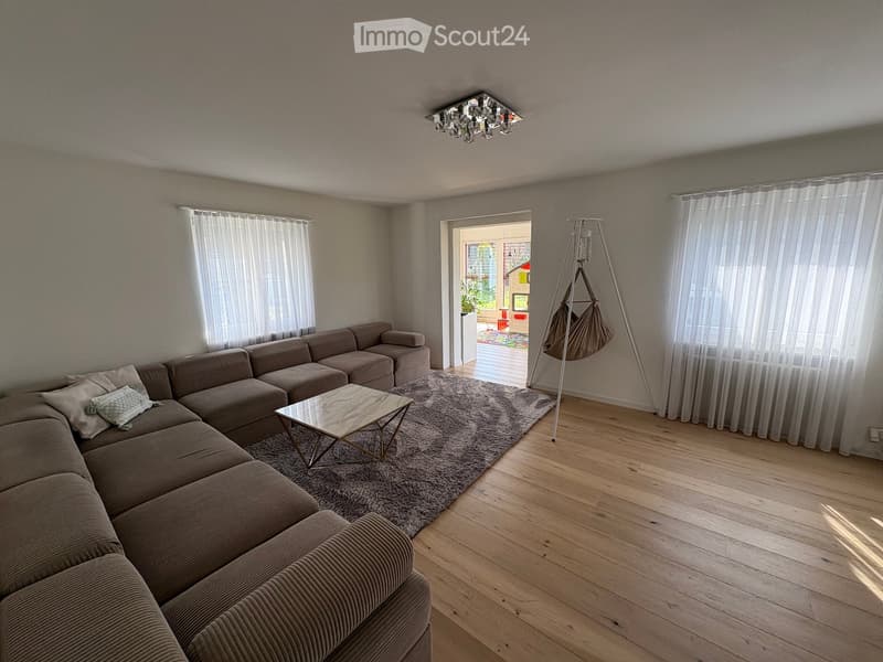 Modern living room with a sectional sofa, coffee table, floor lamp, and large windows with curtains. Wooden flooring and a gray rug.