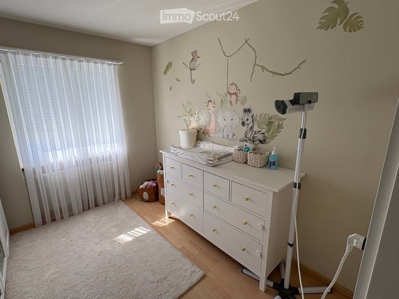 A well-lit nursery with a white dresser, animal-themed wall decals, a large window with white curtains, a soft white rug, and a baby monitor on a stand.