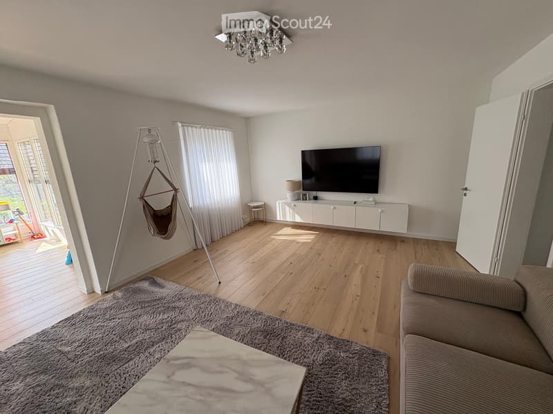 Modern living room with wooden flooring, a grey sofa, a coffee table, a television on a white cabinet, a floor lamp, and a baby chair in the corner.