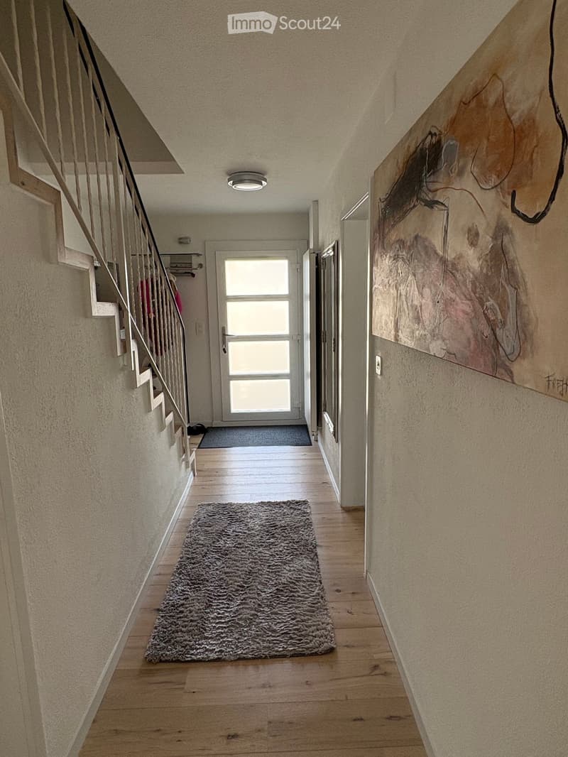 Hallway with wooden flooring, a staircase with metal railing, a large abstract painting on the wall, and a gray shag rug in the center.