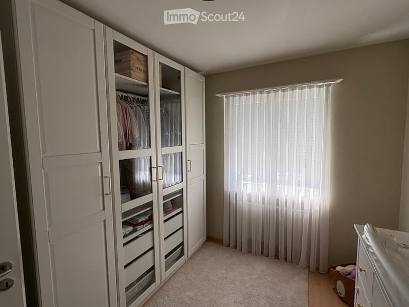 A well-lit bedroom with a large white wardrobe, a neatly made bed, and a curtained window allowing natural light to enter.
