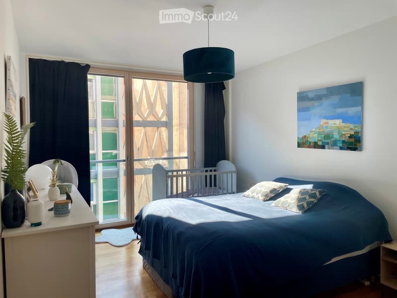Modern bedroom with a large bed, nightstand, and a view of a cityscape through large windows. Features a painting on the wall and decorative items on the nightstand.
