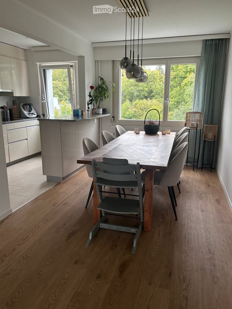 Modern dining area with a wooden dining table and grey chairs, adjacent to a kitchen with white cabinets and stainless steel appliances. Large windows offer a view of greenery, complemented by pendant lights and a plant on the counter.
