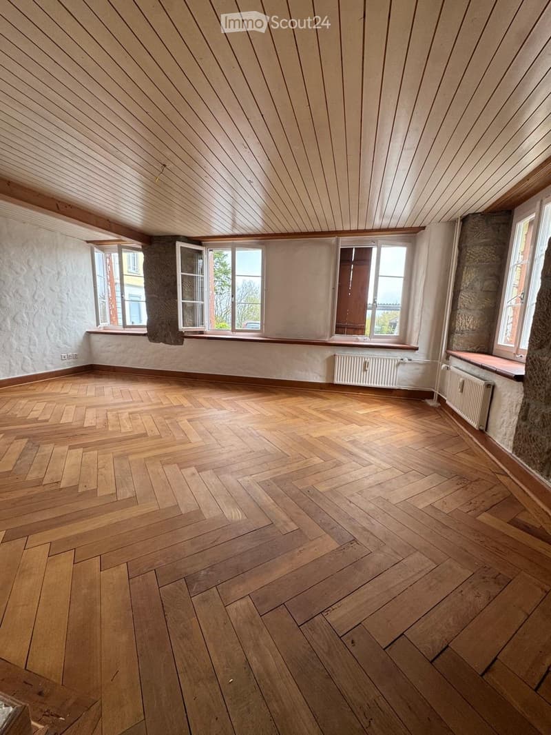 Empty room with wooden herringbone flooring, exposed wooden ceiling beams, and large windows with wooden frames. The walls are painted in a light neutral color, and there are radiators along the walls.