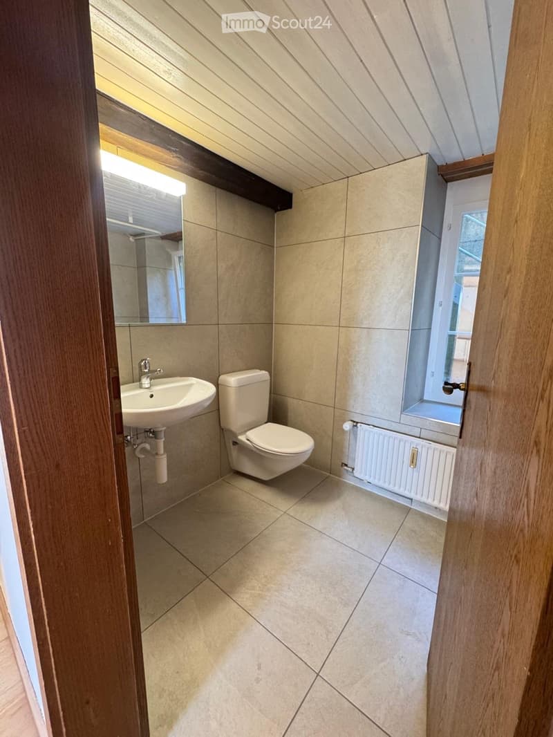 Modern bathroom with beige tiles, a sink, toilet, mirror, and a radiator. The room has a slanted ceiling and a window letting in natural light.