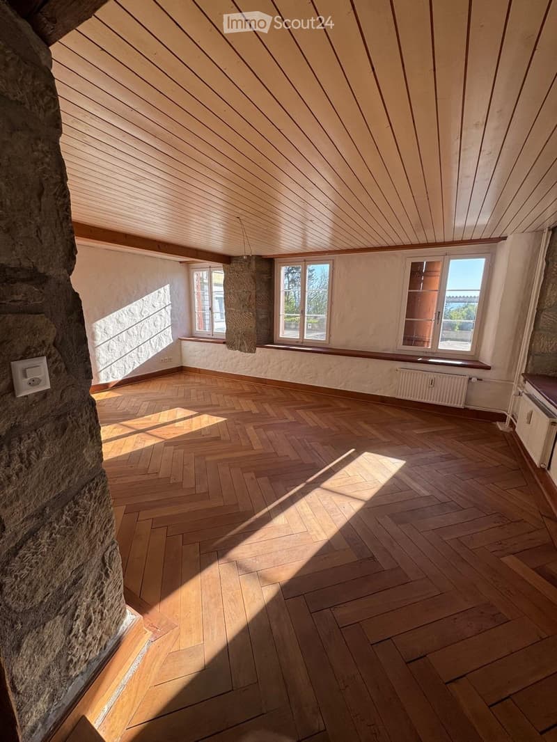 Empty attic room with wooden herringbone flooring, slanted wooden ceiling, and large windows allowing natural light. Exposed stone wall on one side.