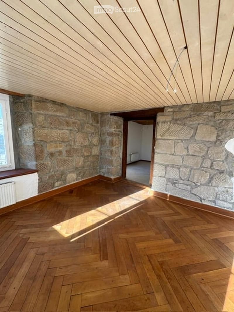 Empty room with exposed stone walls, herringbone wooden flooring, and a slanted wooden ceiling. Natural light streams in from a window on the left.