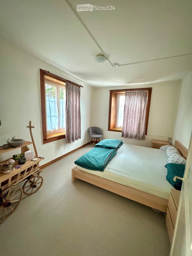 Cozy bedroom with a wooden bed, two windows with curtains, a small wooden table with decorative items, and a simple chair.
