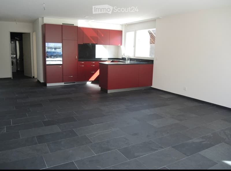 Modern kitchen with red cabinets, black countertops, and dark tiled flooring. Features a central island, sink, and oven.
