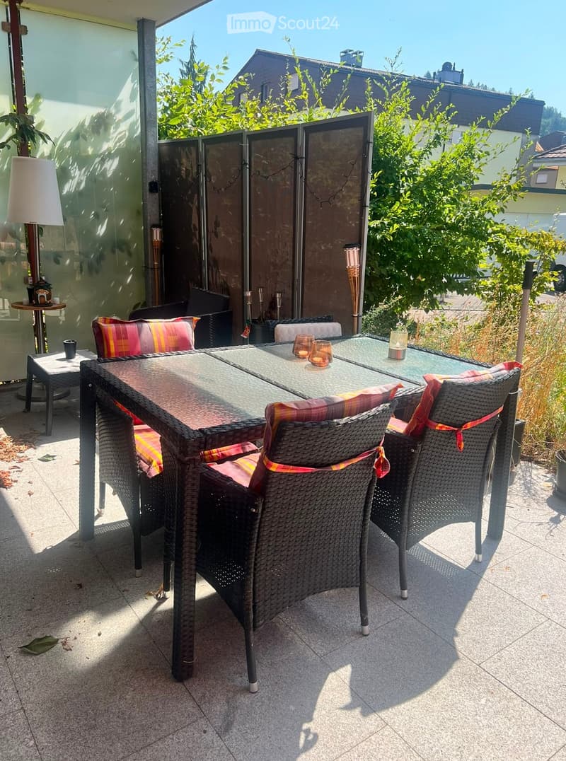 Balcony with a glass table, wicker chairs, colorful cushions, and a small side table with a lamp and cup.