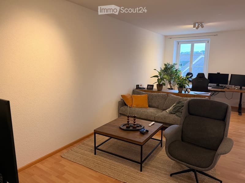 A modern living room with a grey sectional sofa, wooden coffee table, armchair, and a desk with a computer setup. The room features wooden flooring, large windows, and decorative plants.