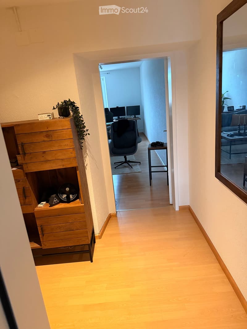 A narrow hallway with light wooden flooring and white walls, leading to an open doorway that reveals a room with a desk, office chair, and computer monitors.