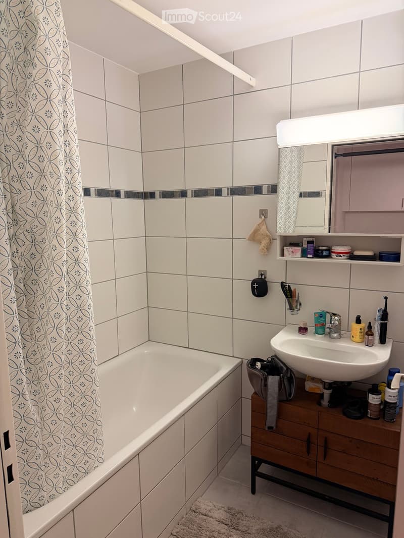 Modern bathroom with white tiled walls, a bathtub, a sink with a mirror, and a wooden cabinet holding various toiletries and a handbag.