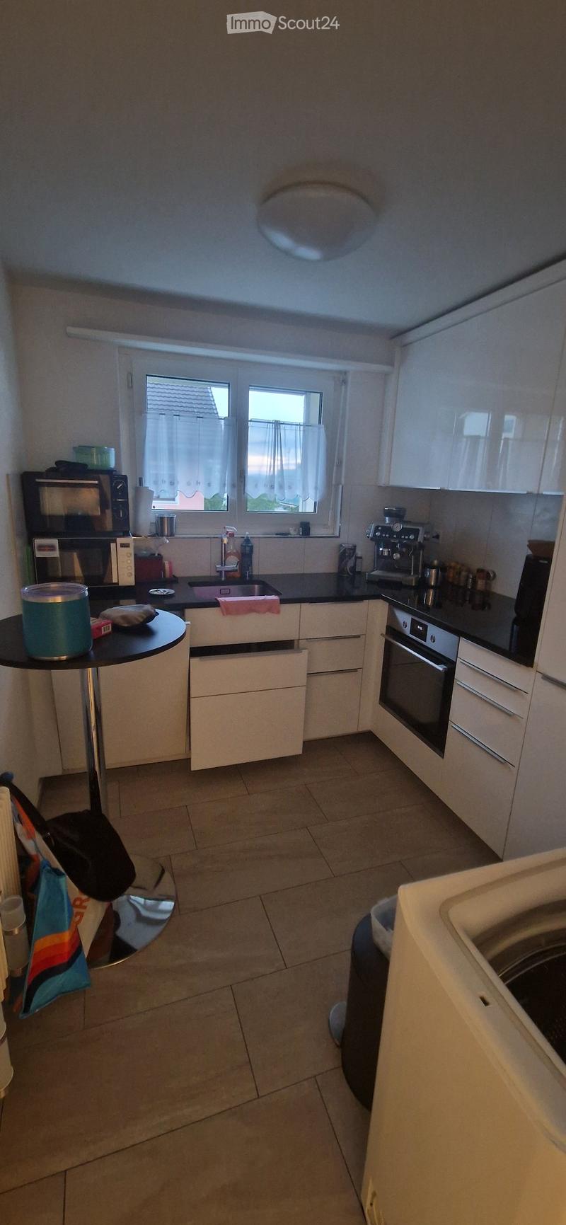 Modern kitchen with white cabinets, black countertops, and stainless steel appliances including a microwave, oven, and coffee maker. Features a sink, faucet, and a small round table with a cup and other items.