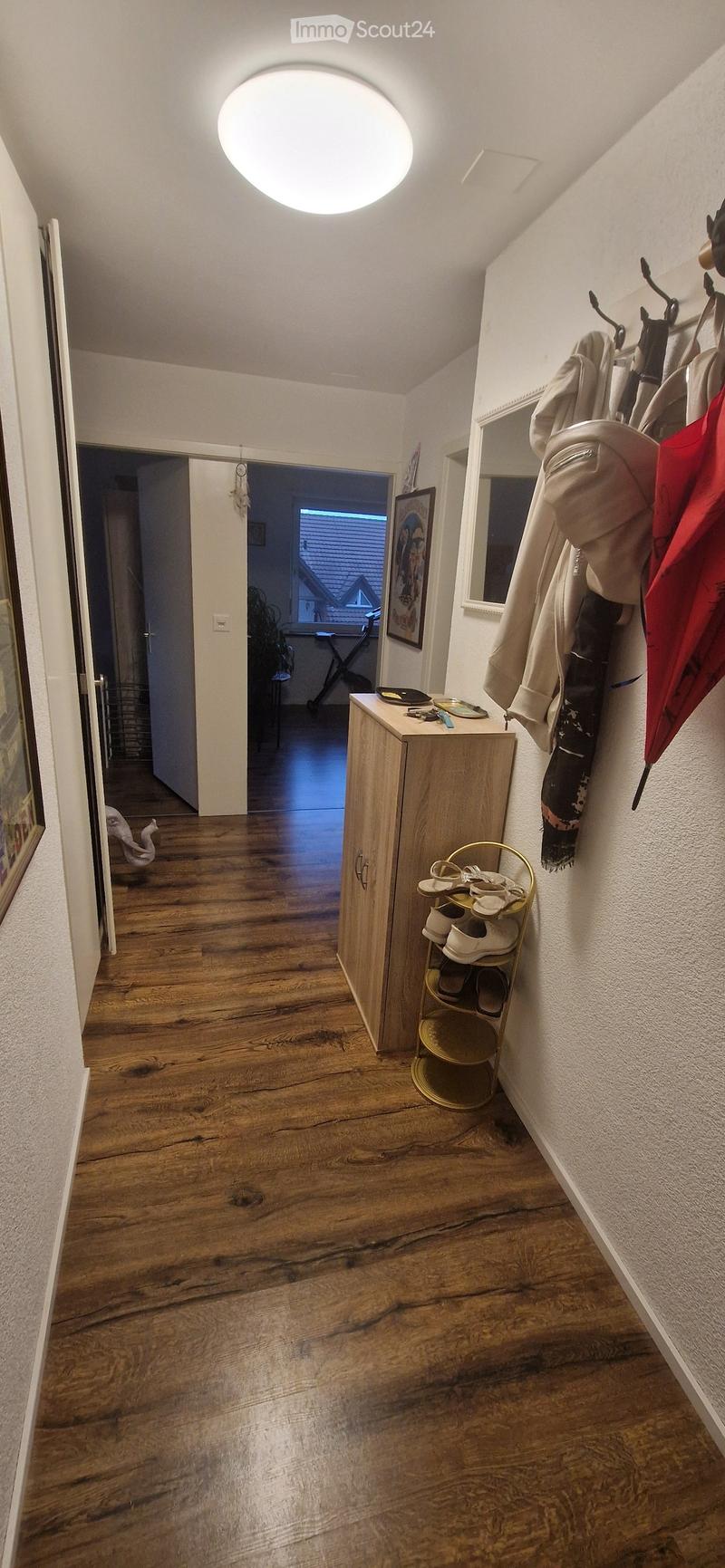 A well-lit hallway with wooden flooring, a coat rack with jackets and shoes, a small wooden cabinet with keys and a mirror, and a round ceiling light.