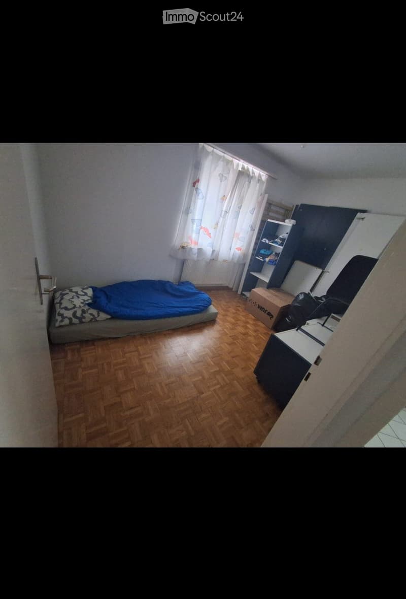A small bedroom with wooden flooring, a bed with a blue blanket, a shelf, a chair, and a desk.