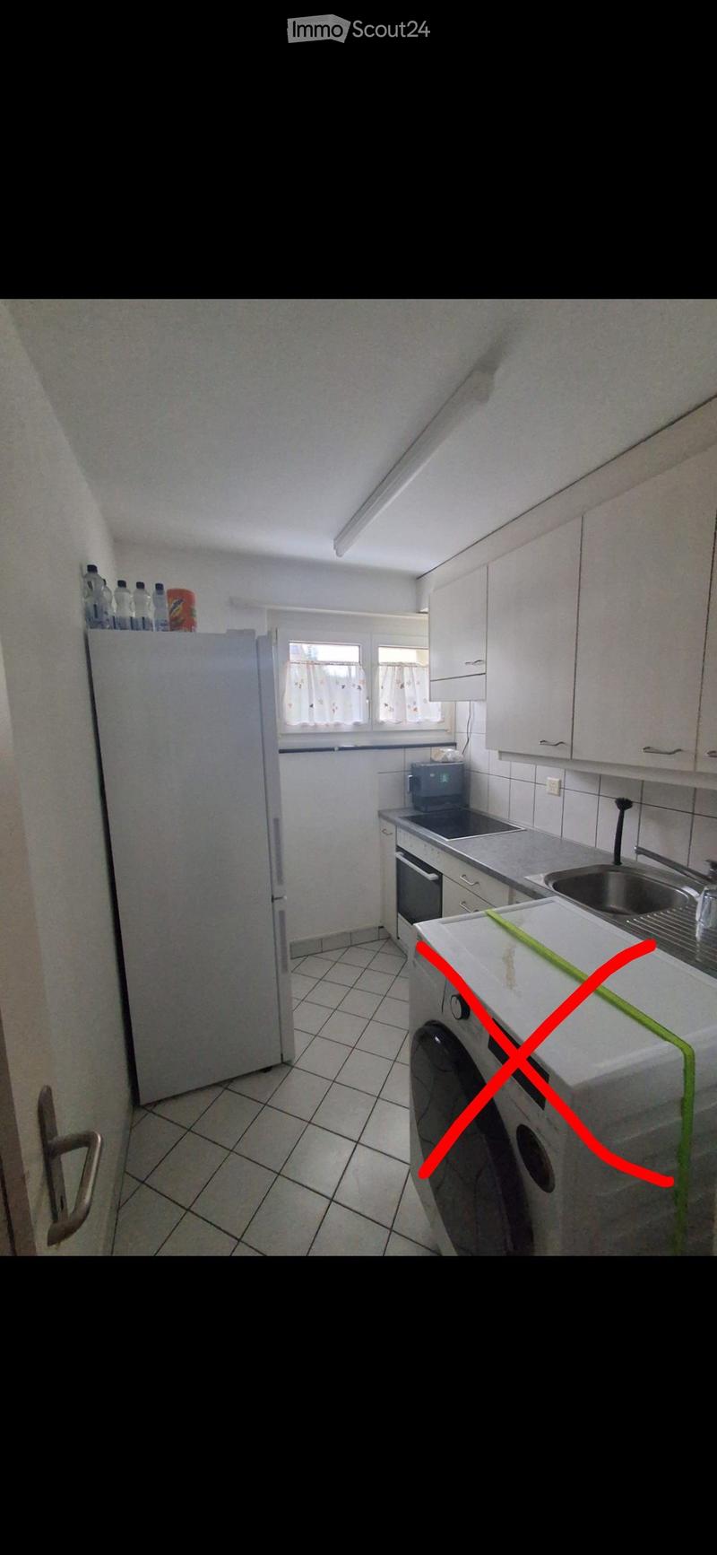 A small kitchen with white cabinets, a refrigerator, microwave, oven, sink, and washing machine. The space is well-lit with natural light from a window.
