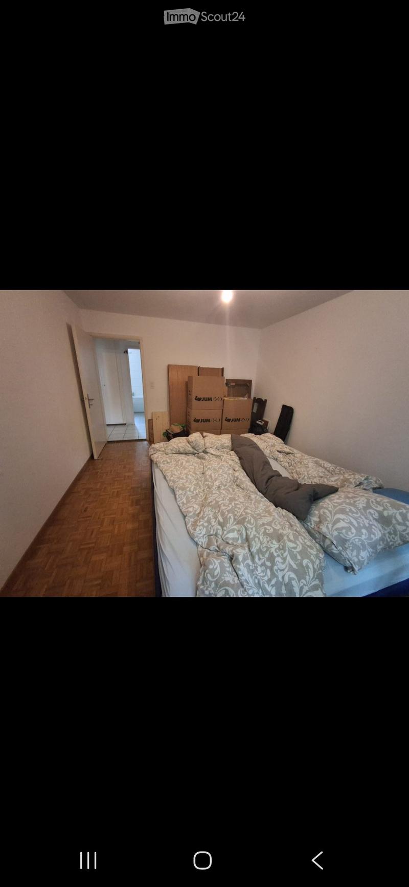 Bedroom with wooden flooring, a bed with patterned bedding, a chair, and several cardboard boxes stacked against the wall.