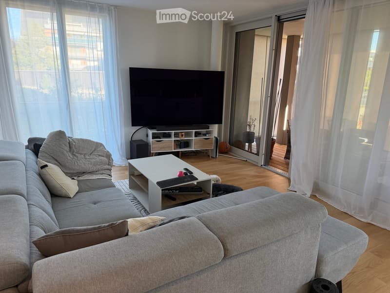 Modern living room with grey sectional sofa, white coffee table, flat-screen TV, and large windows with sheer curtains. The room features wooden flooring and a sliding glass door leading to a balcony.