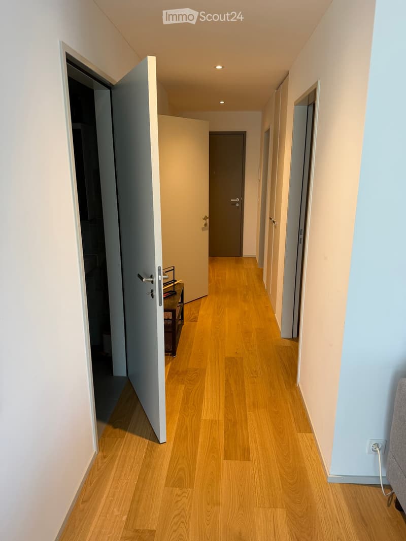 A clean, well-lit hallway with light wooden flooring and neutral-colored walls. Several doors are visible, including an open door on the left leading to another room.