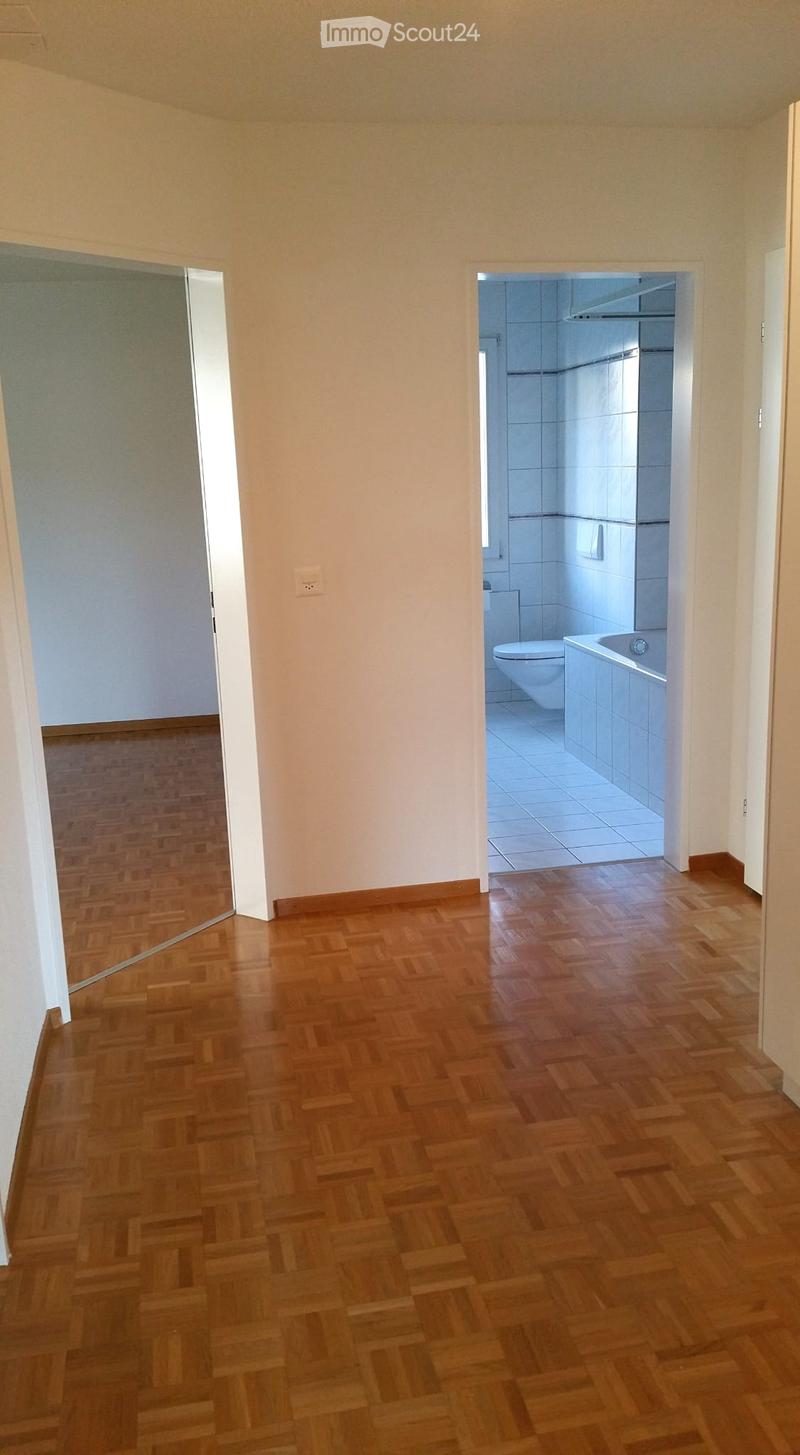 Empty hallway with wooden flooring, leading to a bathroom with white tiles, a bathtub, and a toilet.