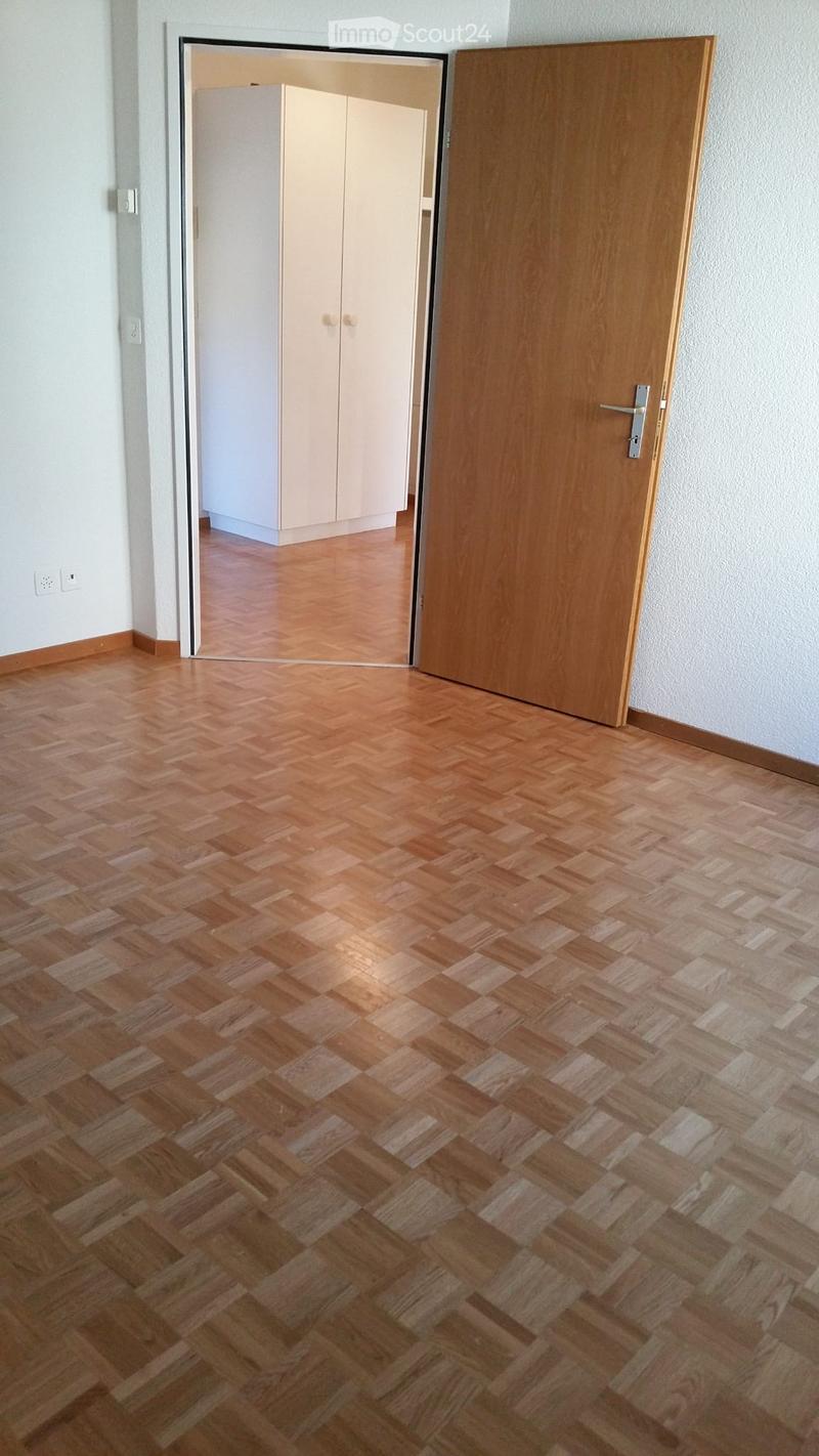 Empty room with wooden herringbone flooring, white walls, and a partially open wooden door leading to another room with white cabinets.