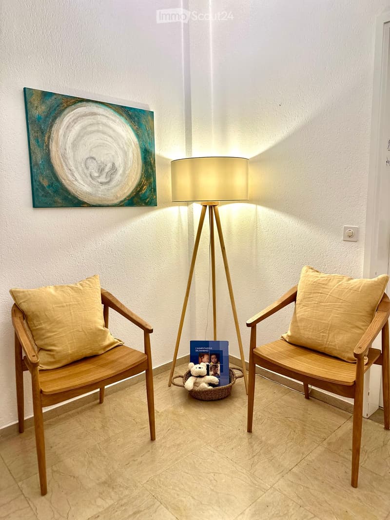 A small, well-lit room with two wooden chairs, a floor lamp, a painting on the wall, a basket with a book and a toy, and a beige cushion on each chair.