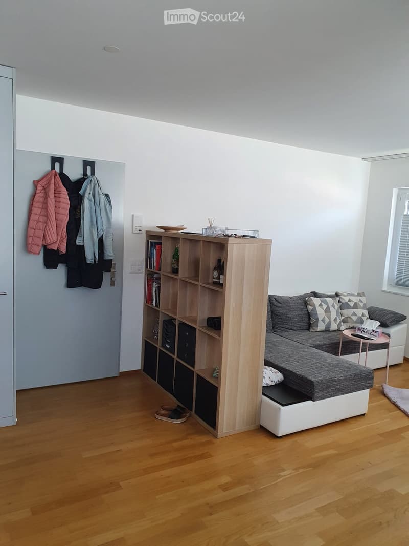 Modern living room with a grey sectional sofa, wooden bookshelf, small round table, and coat rack with jackets.