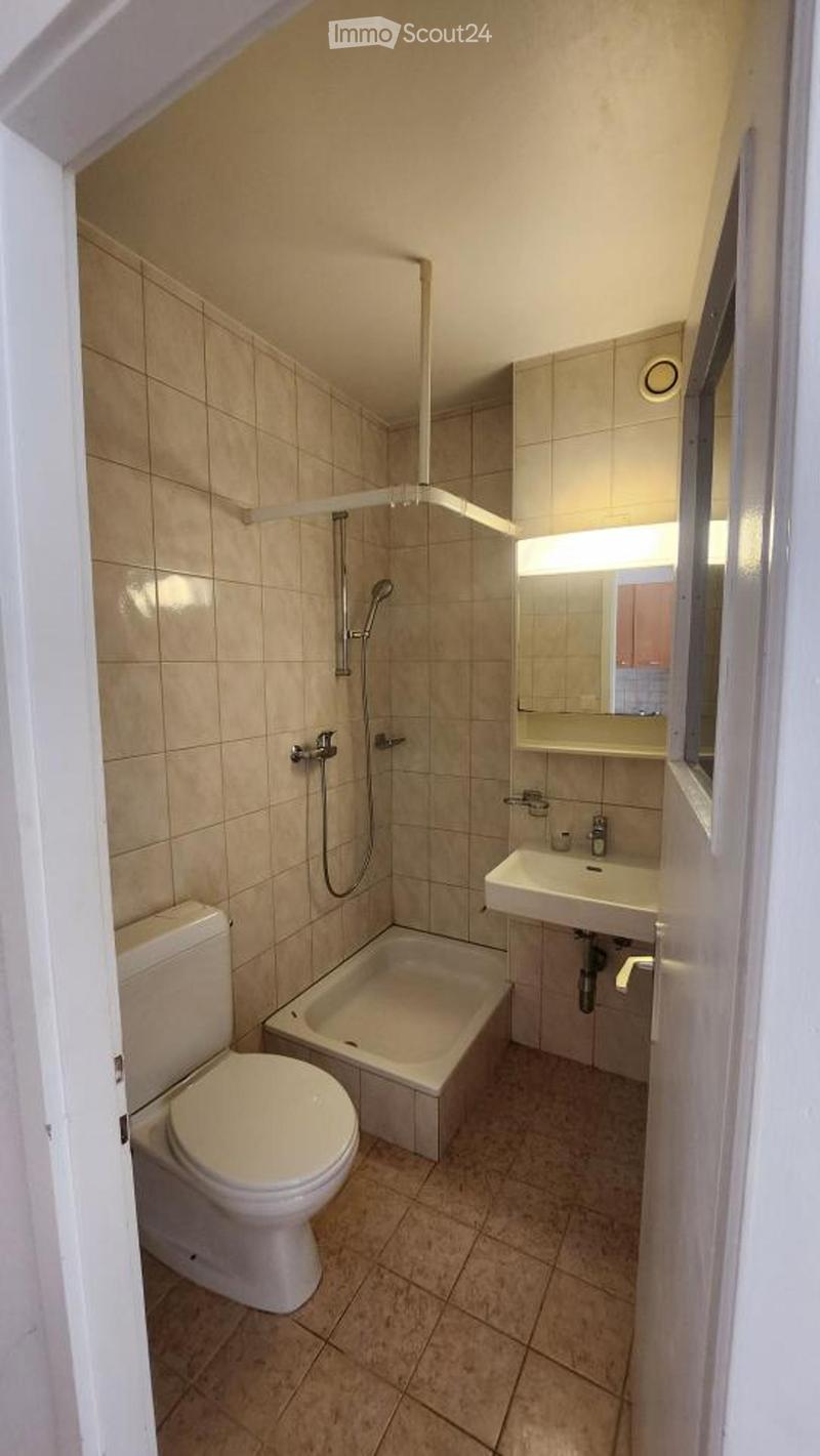 Compact bathroom with beige tiles, a toilet, a bathtub with a showerhead, a sink with a mirror, and a small window.