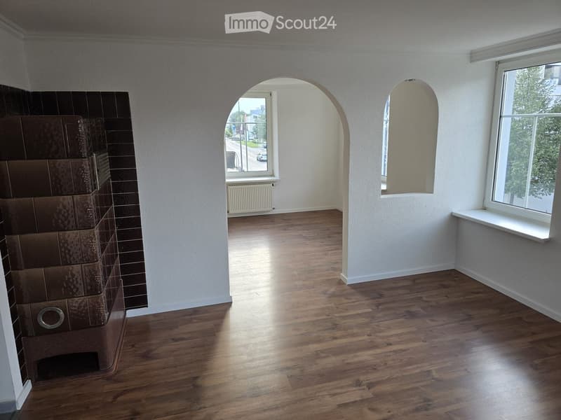 Empty room with wooden flooring, white walls, large windows, and an arched doorway leading to another space.