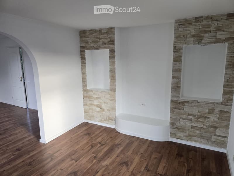 Empty room with wooden flooring, white walls, and a stone accent wall. Features an arched doorway and a window niche.