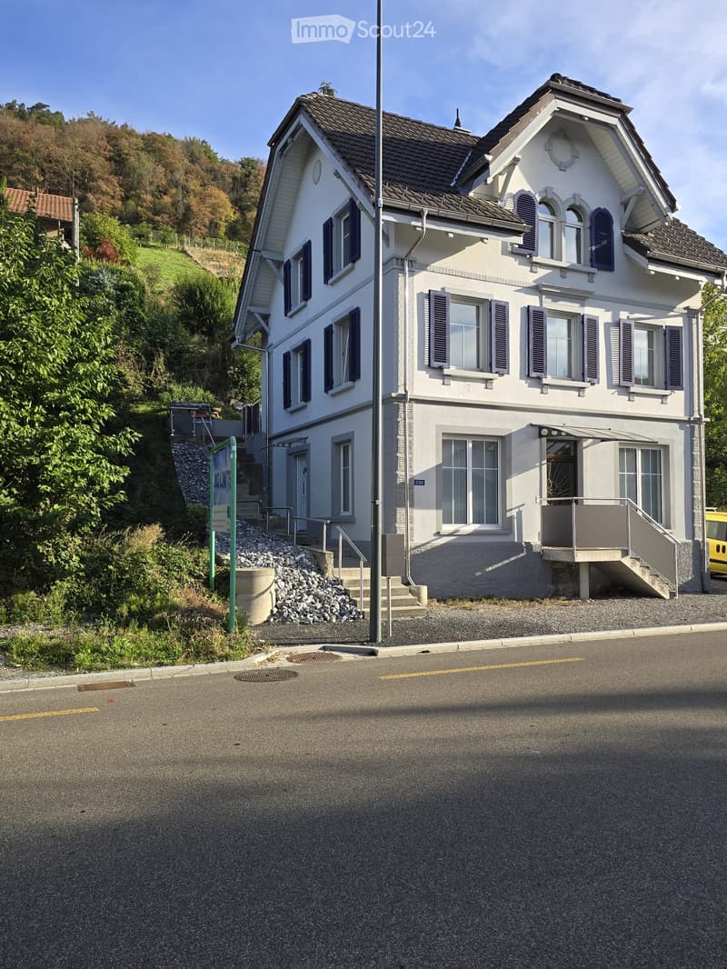 Two-story house with white exterior, multiple windows with shutters, and a small balcony on the first floor. The house is situated on a raised ground floor with steps leading up to it.
