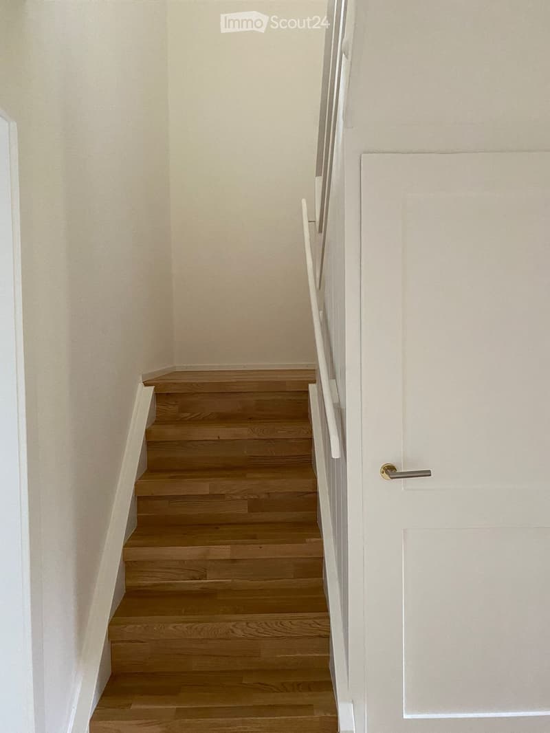 A narrow hallway with wooden stairs leading upwards, white walls, and a closed white door on the right.