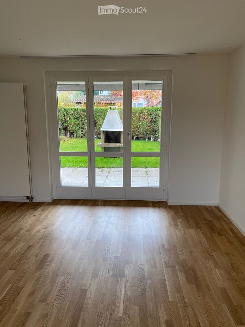 Empty room with wooden flooring and large glass doors leading to an outdoor area with a barbecue and garden.