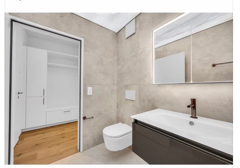 Modern bathroom with a white sink, toilet, and mirrored cabinet. The room features light grey tiled walls and a wooden floor.