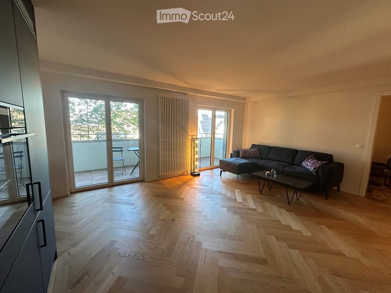 Modern living room with wooden flooring, large sliding glass doors leading to a balcony, a dark grey sectional sofa with patterned cushions, a black coffee table, and a tall floor lamp near the balcony entrance.