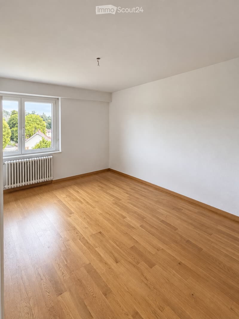 Empty room with wooden flooring, white walls, and a large window with a view of greenery and a house outside. A radiator is visible below the window.