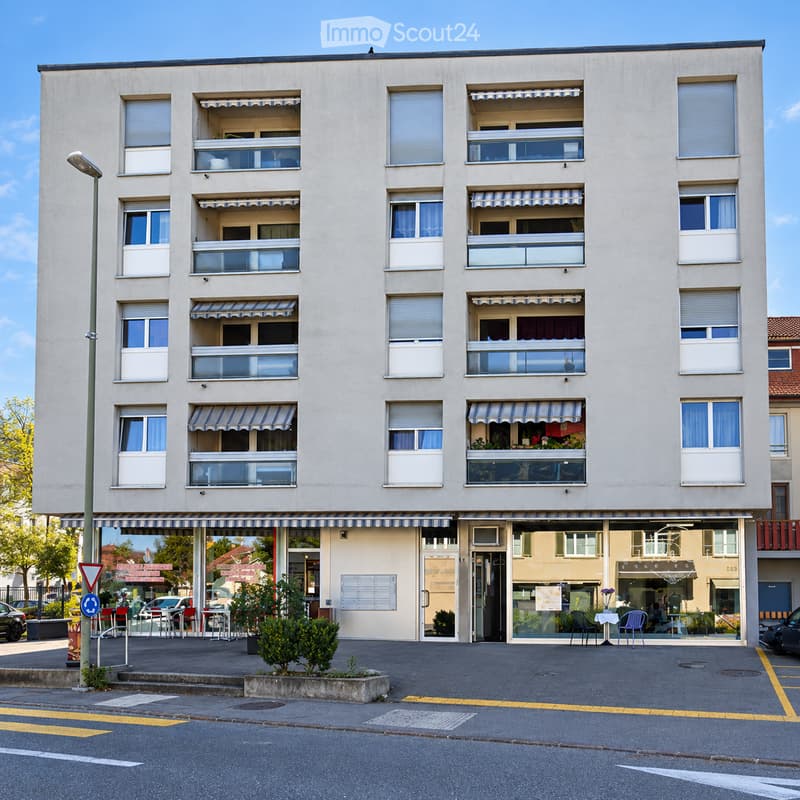 Four-story residential building with multiple balconies, striped awnings, and a storefront at the ground level. Windows have white frames and curtains.