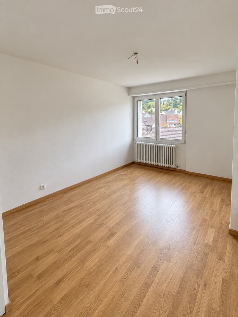 Empty room with light wooden flooring, white walls, and a large window with a radiator below it.