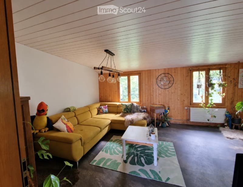 Bright living room with wooden panel walls, large windows, yellow sectional sofa, white coffee table, and decorative plants.