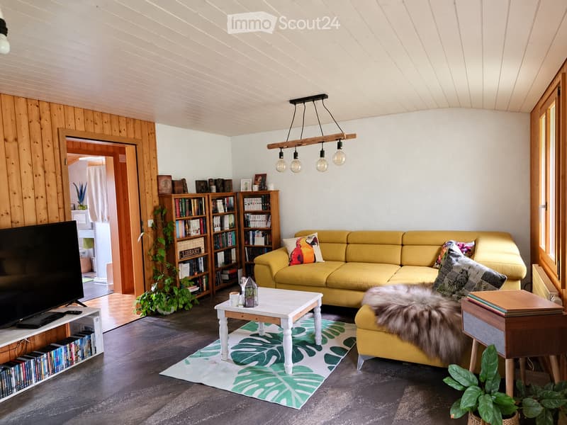 Bright living room with a yellow sofa, white coffee table, bookshelf, and television. Features wooden walls, pendant lighting, and a leafy green rug.
