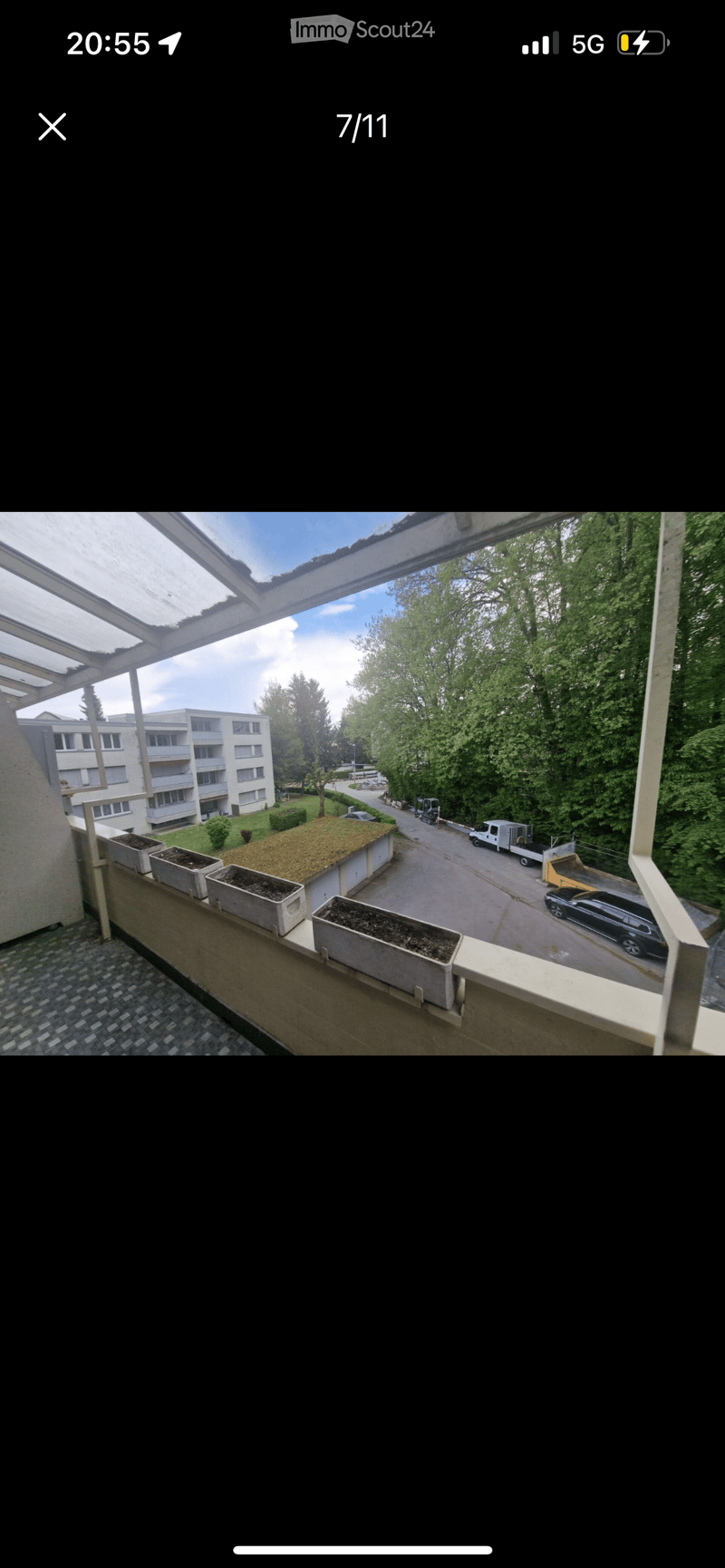 Balcony of an apartment building, with concrete planters, metal railing, asphalt covered ground, green bushes, trees, multi story building, road, parked cars.