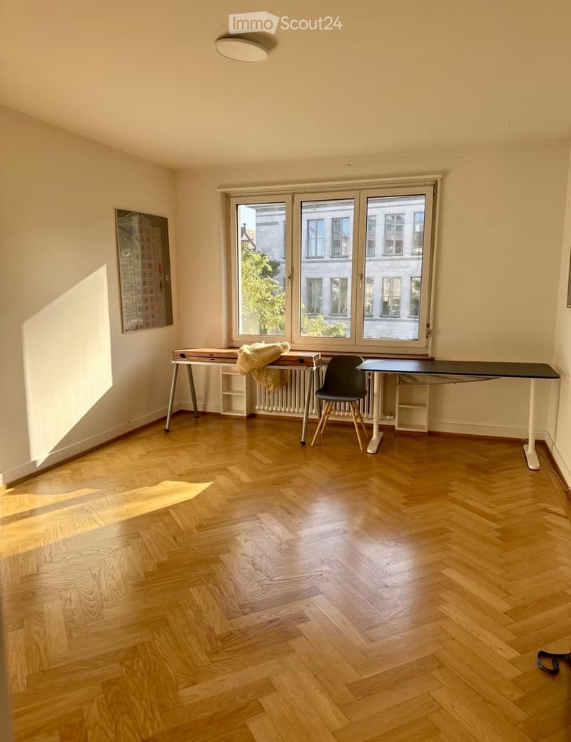 Bright room with wooden herringbone flooring, large windows, a table with a chair, a radiator, and a picture frame on the wall.
