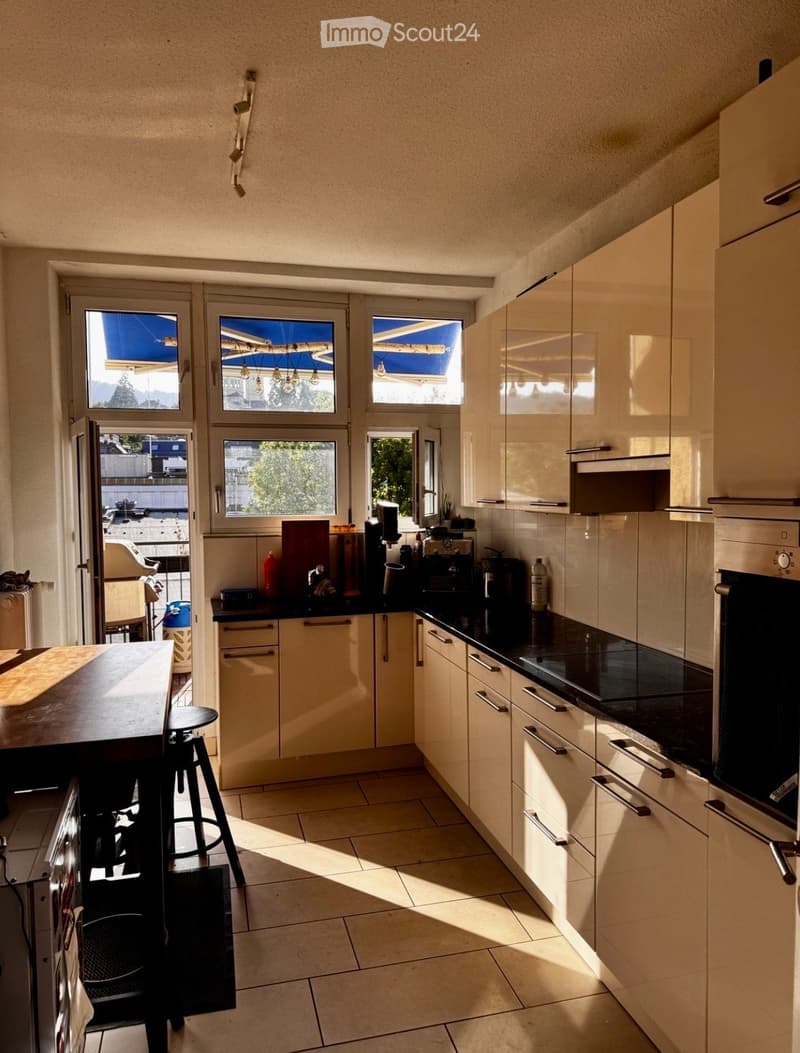 Modern kitchen with light-colored cabinets, black countertops, and a central island with a stool. Appliances include an oven and coffee maker, with a window offering a view of the outside.
